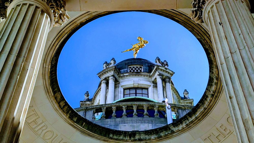Wide shot of the dome of the Bank of England building, with the bronze figure of Ariel, the wind goddess, standing on top against a blue sky. Ariel appears in a dynamic pose with draped fabric flowing around her.