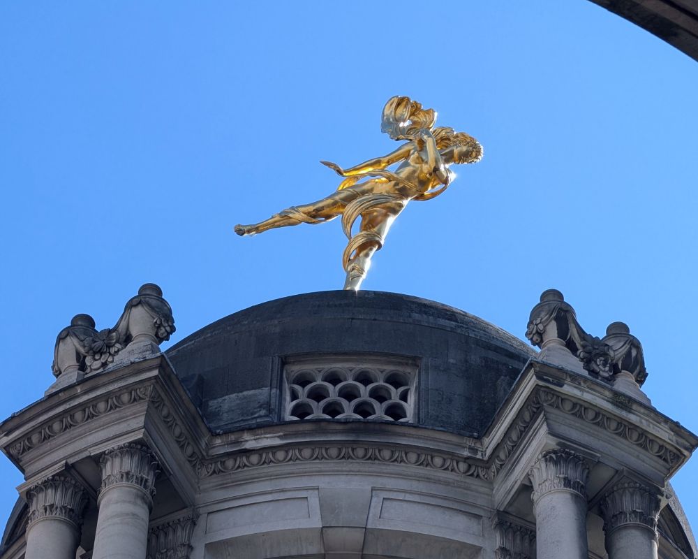 Closer view of the Ariel statue atop the dome, showing more detail of the figure’s posture and the dramatic sweep of her robes. Architectural details of the dome are visible below.

