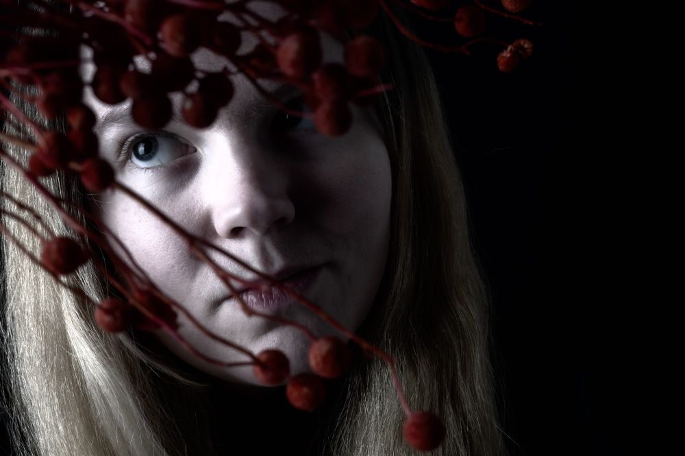A soft-lit portrait of a woman with long blonde hair, gazing slightly upward and to the side. Her face is partially obscured by clusters of red, berry-like dried flowers in the foreground. The background is black, creating a dramatic contrast with her pale skin and the muted red of the blooms. The lighting is moody and atmospheric, highlighting her eye and cheek while the rest of her face fades gently into shadow.