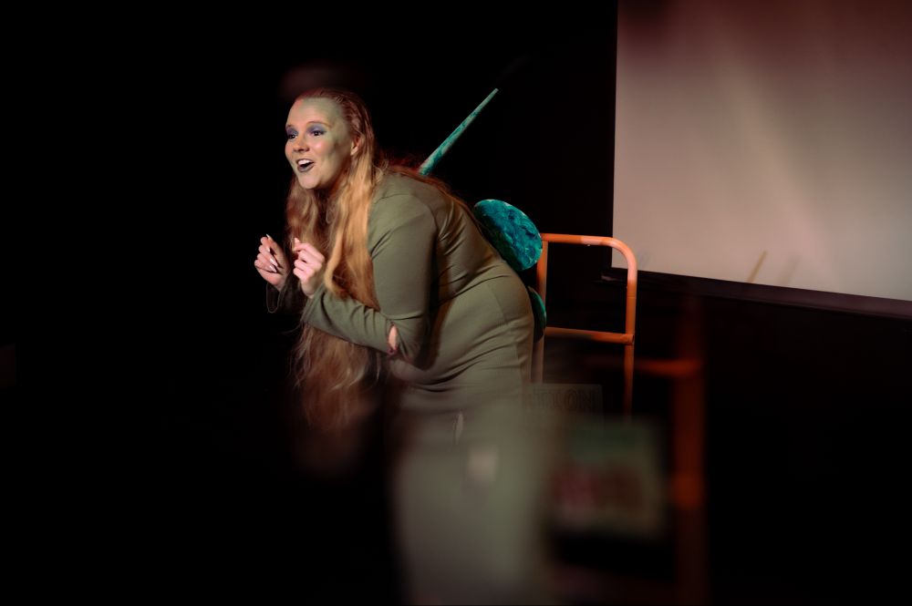 Blonde woman in costume performing on stage in SNOLIGOSTER. Theatrical lighting and surreal in-camera effects create a dreamlike, satirical atmosphere. All images captured live using a Reflect Lens filter.