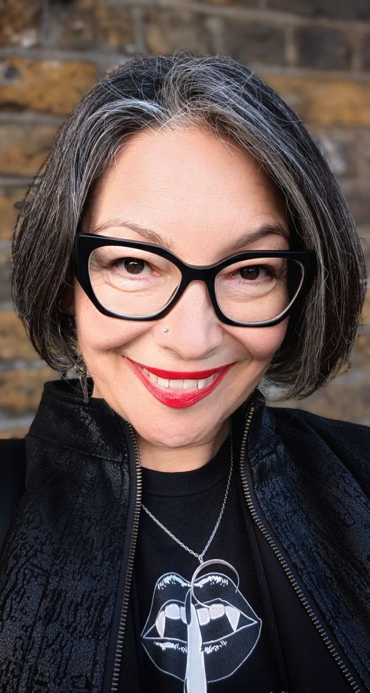 A close-up selfie of a smiling woman with shoulder-length salt-and-pepper hair, wearing bold black Prada glasses. She has a nose stud, bright red lipstick, and is dressed in black with a graphic t-shirt featuring vampire lips and a half moon pendant necklace. Behind her is a brick wall, slightly blurred.
