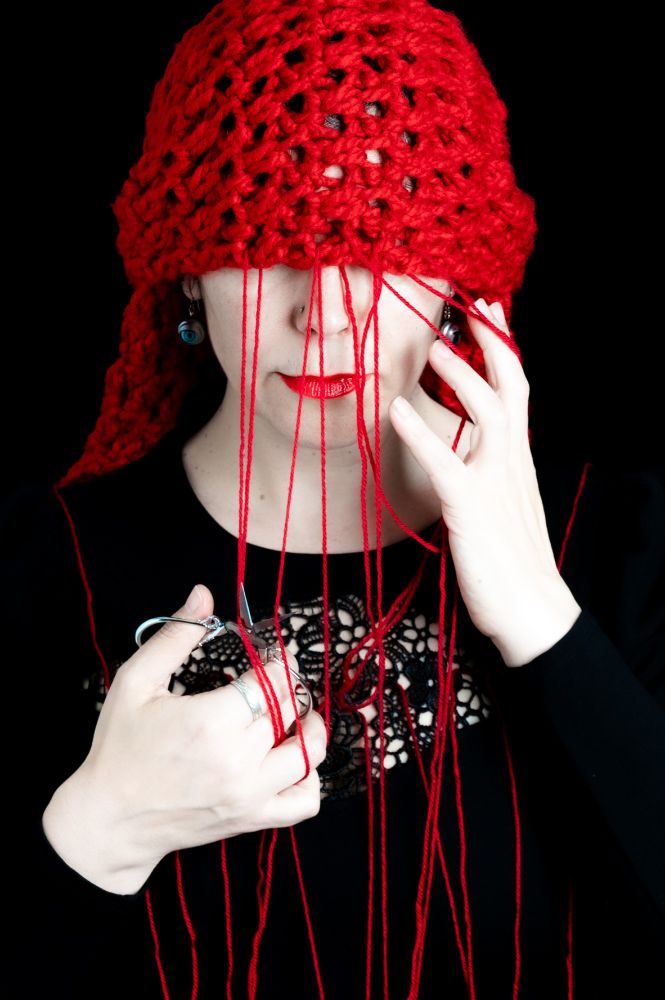 A woman dressed in black holds red yarn threads running from a bright red crocheted head covering down across her face and body. She grips a small pair of ornate embroidery scissors in one hand, poised to cut the threads. Her lips are painted a bold red, mirroring the colour of the yarn. The image references Atropos, the Greek Fate who cuts the thread of life, blending myth, self-portraiture, and historical symbolism.
