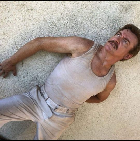 Rhys Darby as Steve Rutherford in Wrecked. He is shot at a Dutch angle as he stands with his back pressed against a white stucco wall. He wears the remains of a cruise ship captain's dress white uniform, which is now just a pair of grubby white trousers and belt and a once-white sleeveless tank top that is doing nothing at all to disguise the fact that he's been doing some significant upper body work at the gym lately. He's rocking the mustache that forever altered my preferences on men's facial hair, and his hair is swept back in a coif that is rather stylish considering his circumstances. He is a bit sunburnt and sweaty and covered in just enough smudges of dirt to look adventurous instead of untidy. He is looking off to one side with an expression of concern, exhaustion, and imminent adrenaline crash and seems quite unaware that this is the hottest photo that will ever be taken of him.