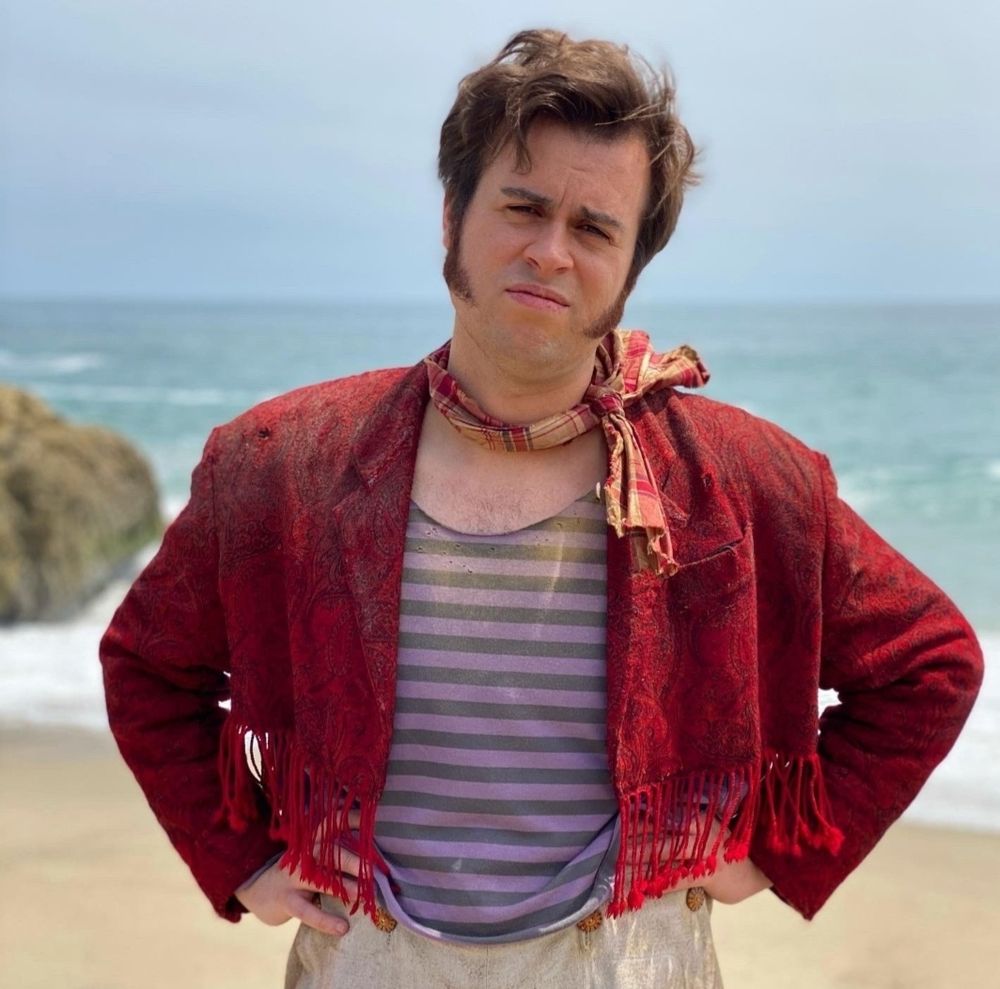 Lucius Spriggs standing on a beach. He wears his fringed red jacket, striped shirt, and that kicky little scarf. His hands are on his hips and he is looking into the camera with a look of disdain and/or annoyance. (This is a BTS shot so the attitude is actually coming from Nathan, but the end result is the same. )