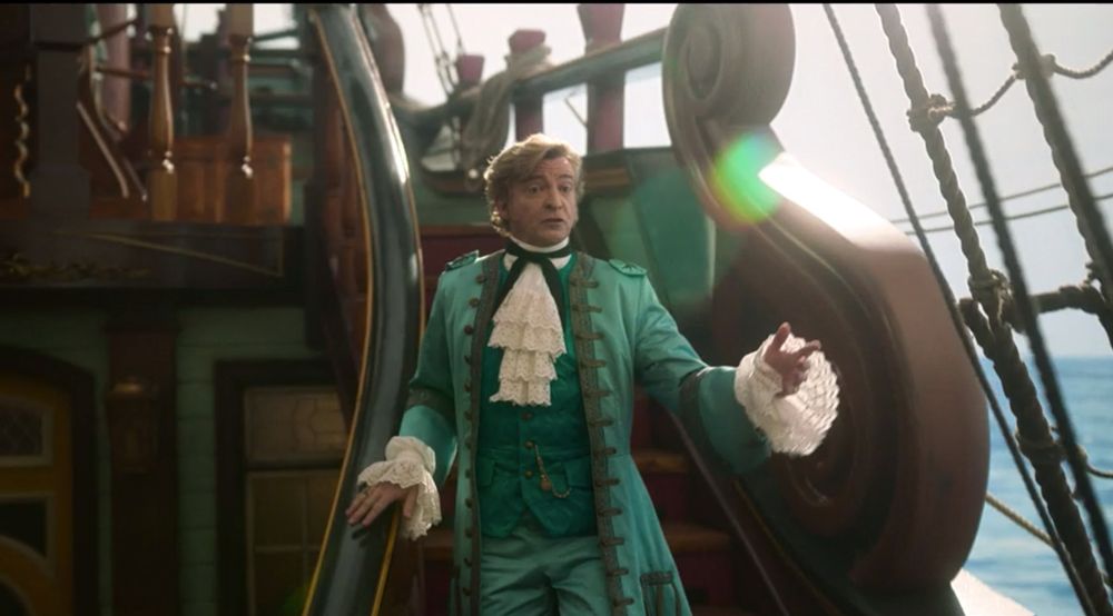 Rhys Darby as Stede Bonnet in S1E1 of Our Flag Means Death. He is walking down the deck staircase of the Revenge during his opening speech. He is wearing his turquoise silk suit with lace jabot and sleeves, and director Taika Waititi has conveniently arranged for a rainbow lens flare to happen right beside his head.
