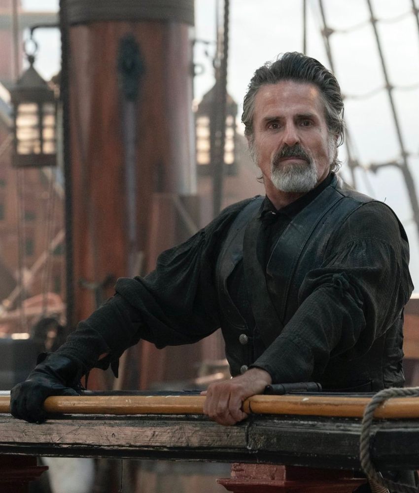 Izzy Hands on the deck of the Revenge, his hands braced on the rail. He wears a black shirt & cravat, black leather waistcoat, black baldric, and one black glove. His black leather pants are not visible in this photo, but rest assured that he is wearing the living fuck out of those as well. He is looking directly at the camera. This is a BTS shot, likely taken after Izzy fanboys all over Blackbeard after Ed suggests that he could take over as captain after Ed kills Stede, a scene that clearly illustrates how Izzy's devotion to Ed & the Blackbeard persona has clouded his ability to anticipate the fact that neurodivergent love will soon upend life at sea as he knows it and introduce him to the transformative powers of eye makeup.