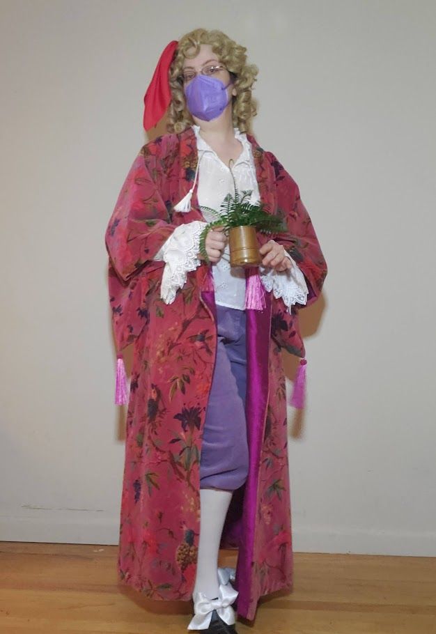 My Stede cosplay from ECCC 2022. I am wearing a pink velvet breakup robe, a white shirt with ruffled sleeves, purple breeches, white stockings, and black shoes with large white satin bows. My blonde curly wig is pretty mid, but hey, someone spent half their budget on pink velvet fabric. I am wearing Ed's red silk as a hair ornament. I am holding a very excellent reproduction of the plant from the fishing boat raid: a rather rangey-looking faux fern in a weathered copper cup.