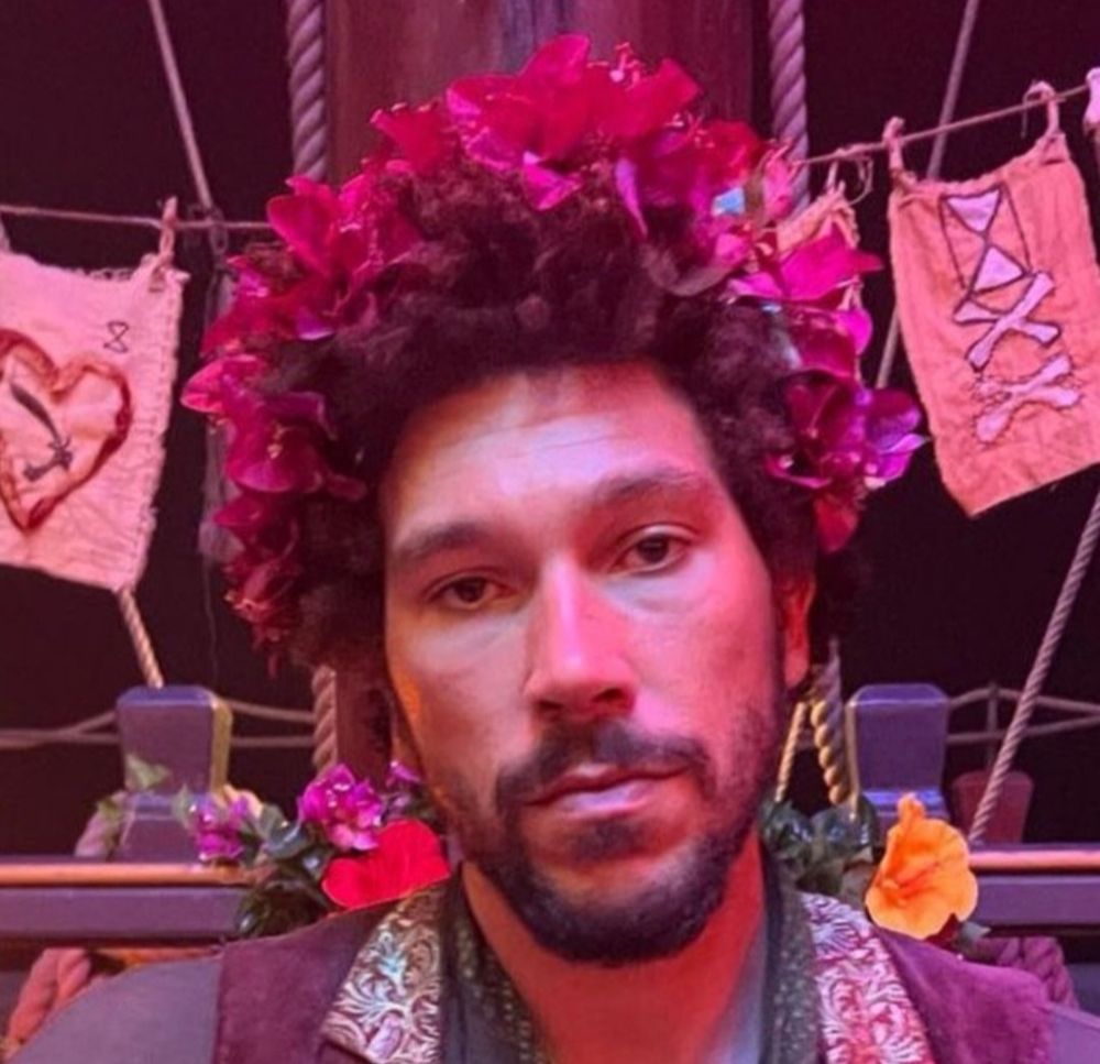 Joel Fry dressed as Frenchie in the Calypso's Birthday episode. He wears a colorful brocade vest and a crown of purply-pink flowers. More flowers and hand-sewn decorative flags are in the background. He is looking directly into the camera with a blank, unamused expression.