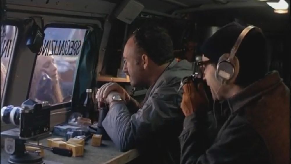 A still from the movie “The Conversation.” We’re inside a van, and through a window you can see a woman applying blush with a brush, while inside the van two men look on sort of lecherously. One of the men is taking a photo of the woman with a camera. The other man is played by the late Gene Hackman.
