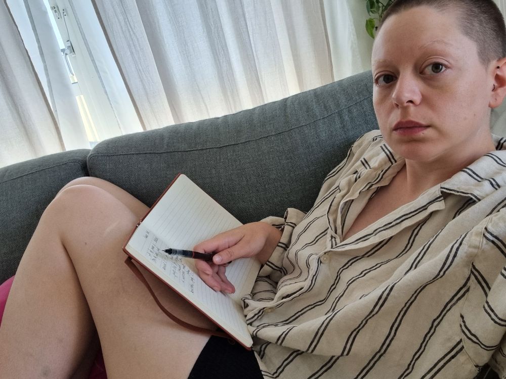 billie sitting on sofa wearing the loosest linen shirt they could find with bike shorts, trying to write in a notebook