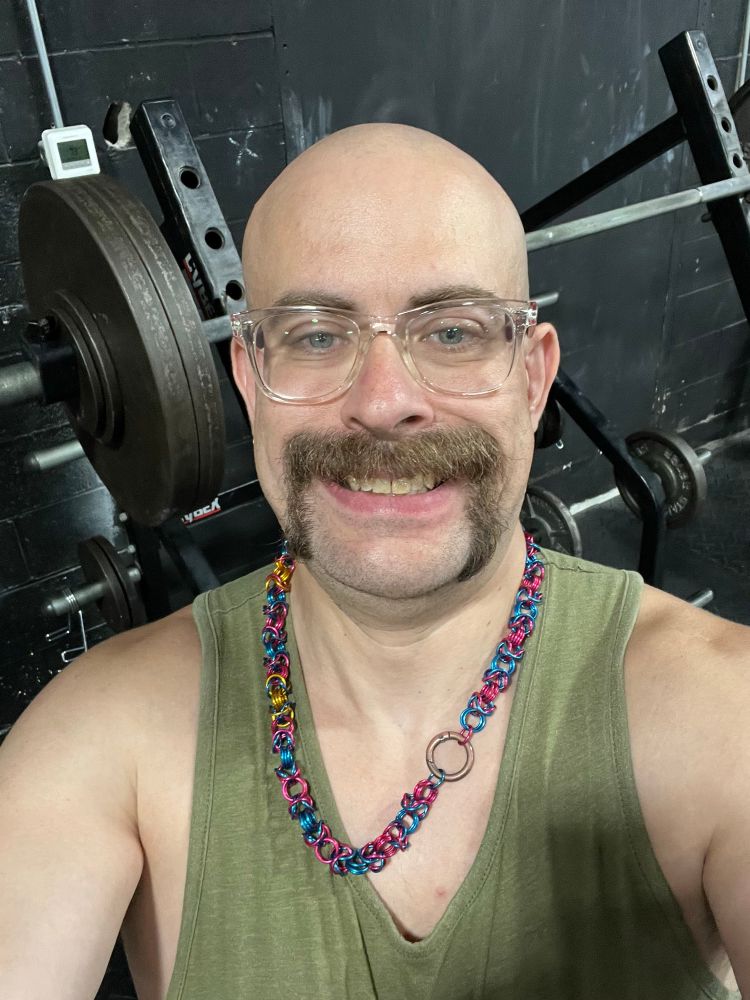 A bald mustached white man in front of a squat rack with a barbell with two 45 pound plates, a 5 pound plate, and a 2.5 pound plate on one end.