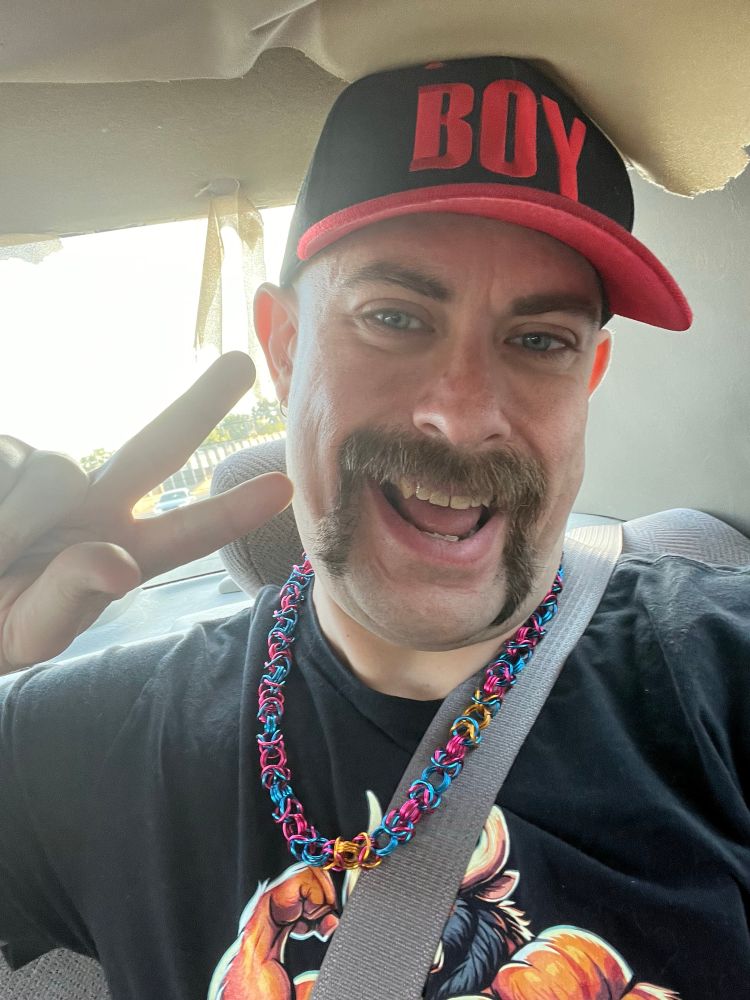 A mustached white man in a hat that says “BOY” doing a peace sign in a car.