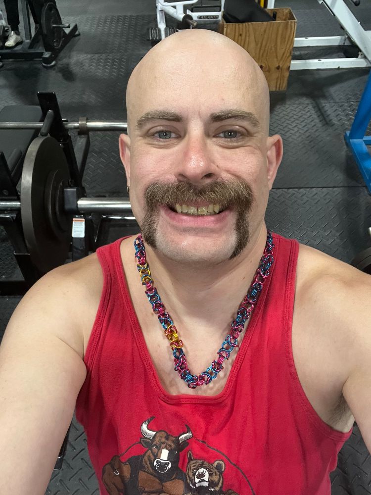 A bald mustached white man in front of a bench press rack with a barbell with a 45 pound plate, a 10 pound plate, and a 2.5 pound plate on one end.
