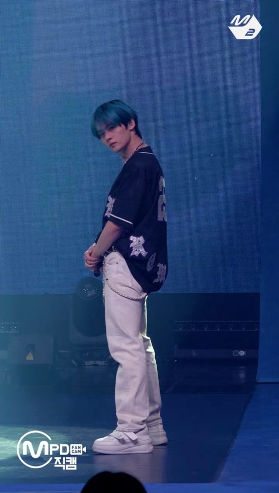 Lee Know on stage wearing a black athletic jersey and white pants. His arms are crossed in front of his pelvis. His hair is blue. 
