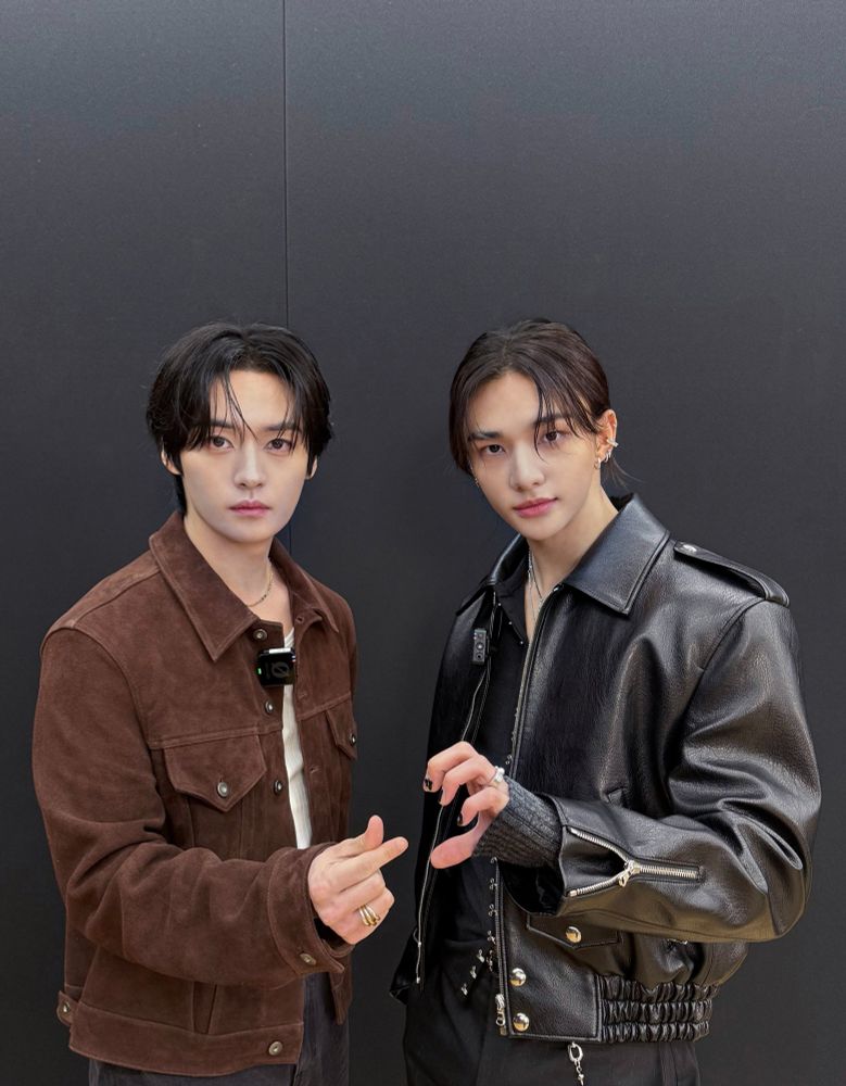 Stray Kids members Lee Know and Hyunjin standing in front of a black wall. Lee Know is wearing a brown jacket with a white shirt and black pants, and Hyunjin is wearing a black leather jacket with a black shirt and black pants. They are holding up two different hearts—Lee Know holding a finger heart, and Hyunjin half a hand heart.