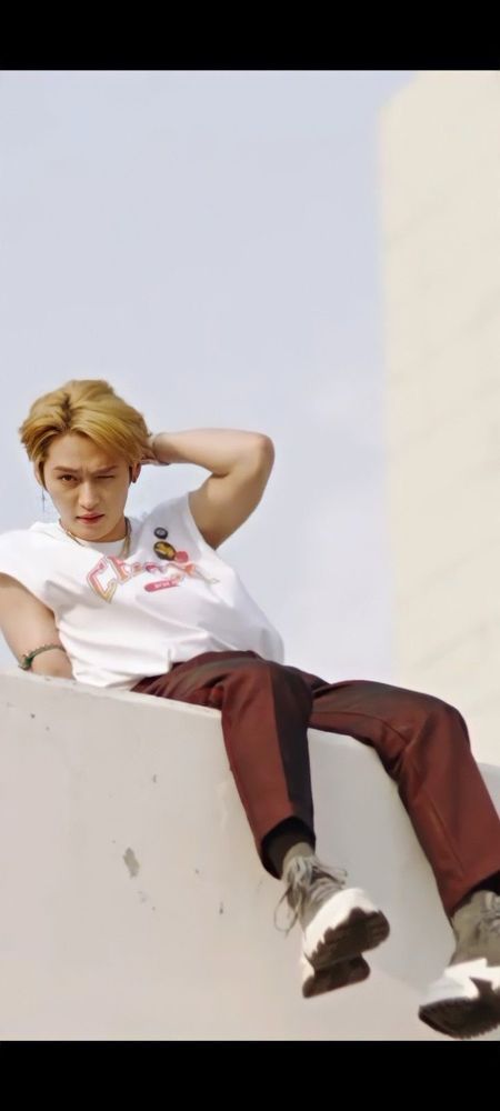 Stray Kids member Lee Know in the “CHEESE” music video. He’s sitting on top of a concrete building wearing a white sleeveless shirt and dark red pants. There’s an accessory around his upper arm accentuating his muscles. He’s leaning back slightly with one hand behind his head. His hair is a brassy blond. 