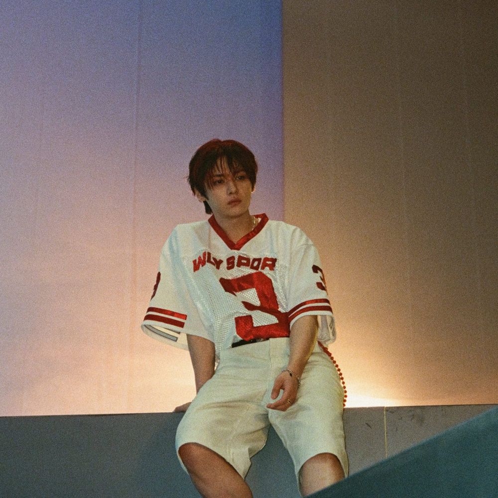 Lee Know posing for a KARMA photo shoot concept. He’s sitting on a wall and wearing an off white and burgundy football jersey.