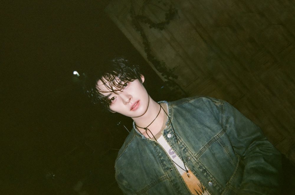 Stray Kids member Lee Know is standing outside wearing a blue-green denim jacket over a graphic tee and a necklace. The picture is taken at an angle so that Lee Know is tilted to the left. 