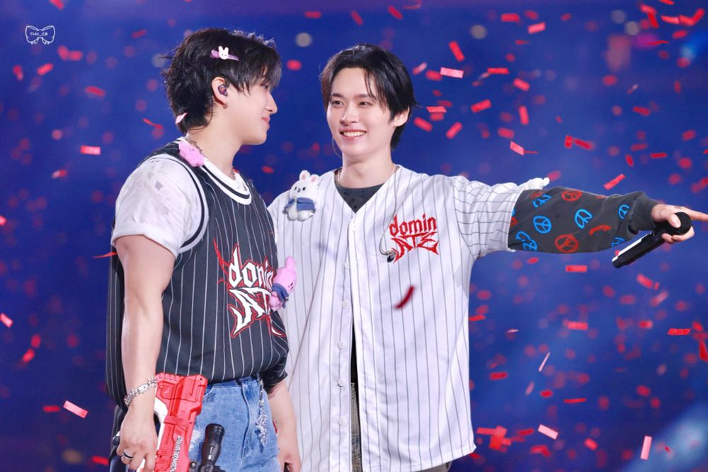 Stray Kids members Lee Know and Changbin on stage at dominATE LA. Lee Know is wearing a tour baseball jersey over a long sleeved black shirt, and Changbin is wearing a tour basketball jersey over a white shirt. Lee Know is showing Changbin something that he’s pointing to off camera (probably the audience) and looking at Changbin with a smile on his face. They’re both holding red water guns. Red confetti is falling around them.