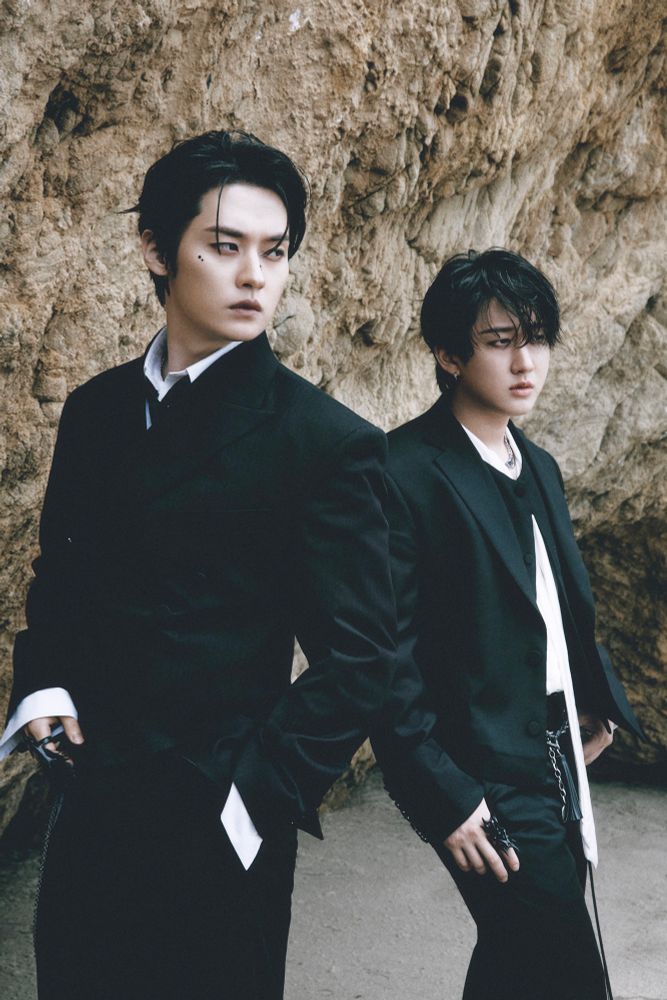 Lee Know and Changbin standing almost perpendicularly with their shoulders touching. They’re both dressed in crisp black suits with white shirts. They’re standing in front of a rocky cliff formation and looking out to the shore off camera with serious expressions.