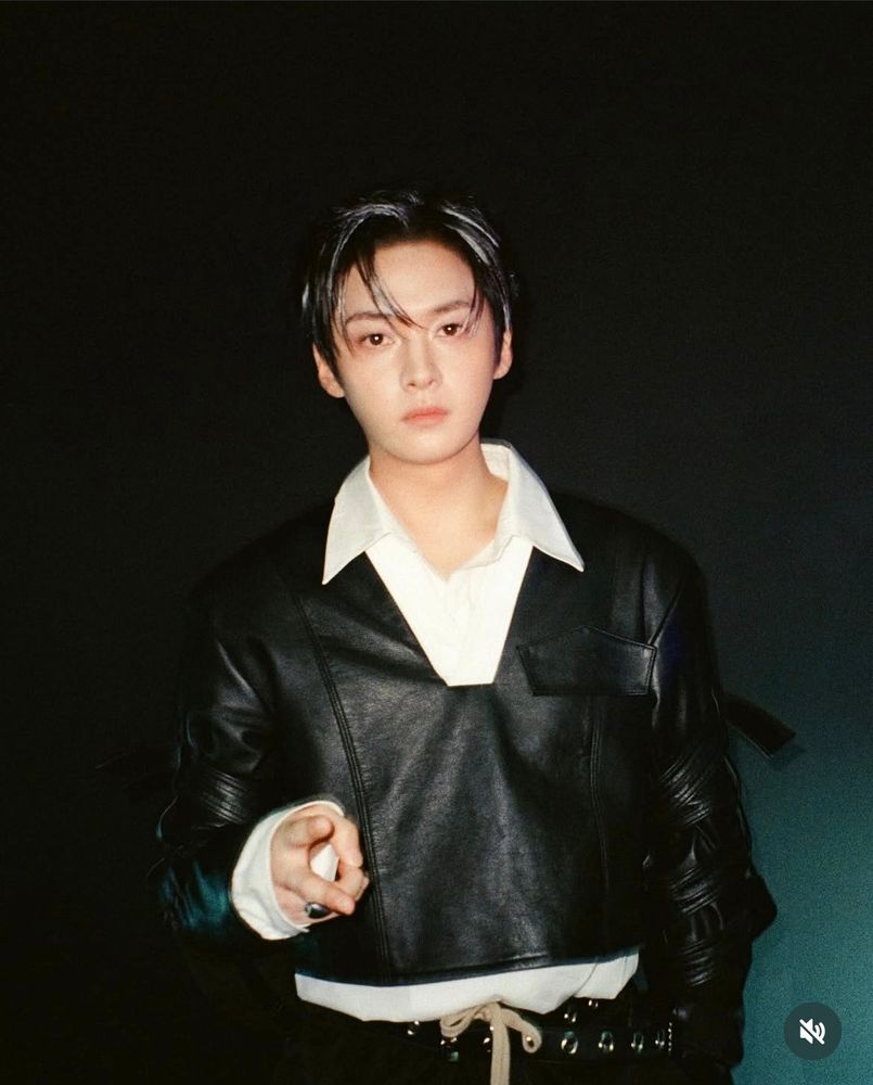 Stray Kids member Lee Know. He’s wearing a black long sleeve leather crop top over a white dress shirt and black pants. He’s holding a peace sign. 
