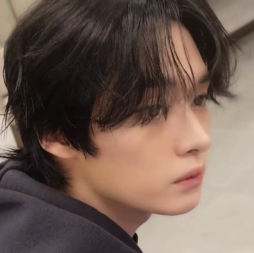 Stray Kids member Lee Know from the neck up looking over his right shoulder at something off camera. He’s wearing a black hoodie with his bangs in his eyes. 