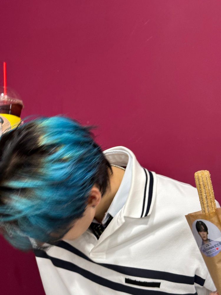 Lee Know holding a churro in one hand and some kind of iced drink (maybe americano?) in the other. The churro packaging and the drink’s cup sleeve have a picture of his face on it. He’s looking down so you can only see the top of his head. The wall behind him is a reddish color.