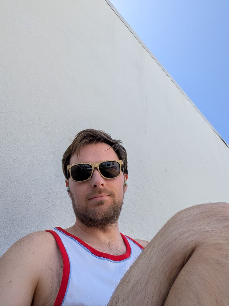 A selfie of me in a tank top showing some leg on the roof wearing sunglasses with a white backdrop and a clear sky peaking out in the corner 