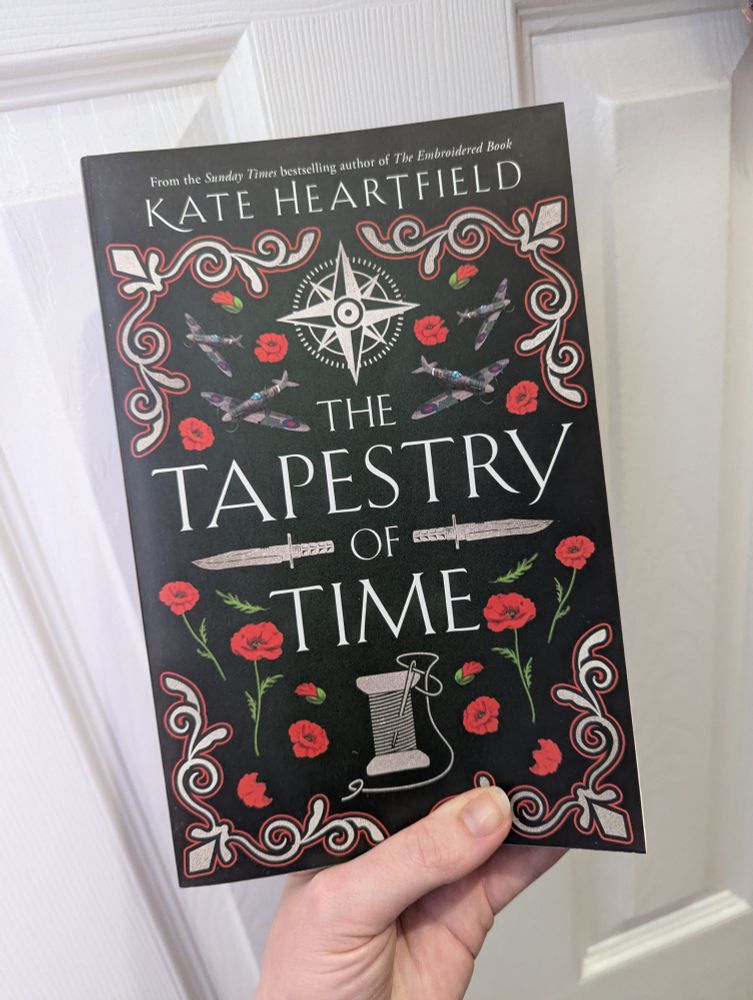 A paperback copy of The Tapestry of Time by Kate Heartfield. The cover features poppies, warplanes, swords and a needle and thread. 