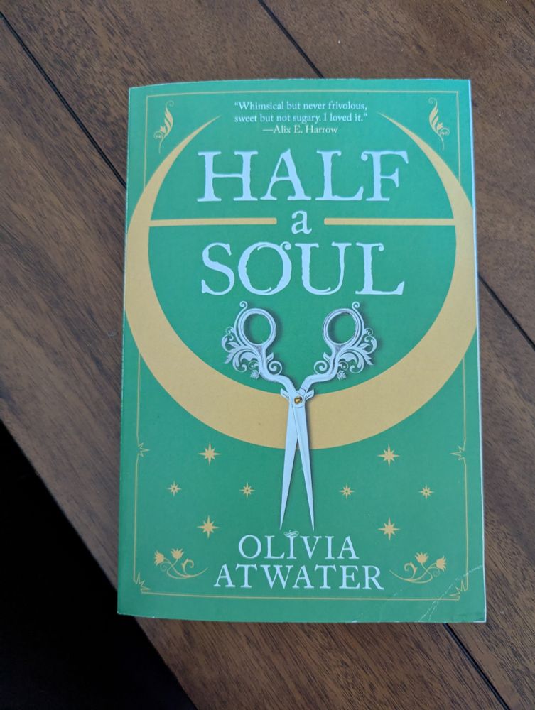 A paperback copy of Half A Soul by Olivia Atwater on top of a dark wooden table. The cover is green with a pair of scissors and a yellow crescent 