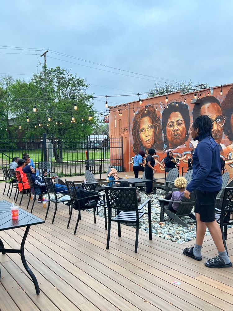 Students sit on dark gray metal lawn chairs placed around a deck and rock pit beside a building. The side of the building has a painted mural depicting several faces on it. String lights hang overhead.
