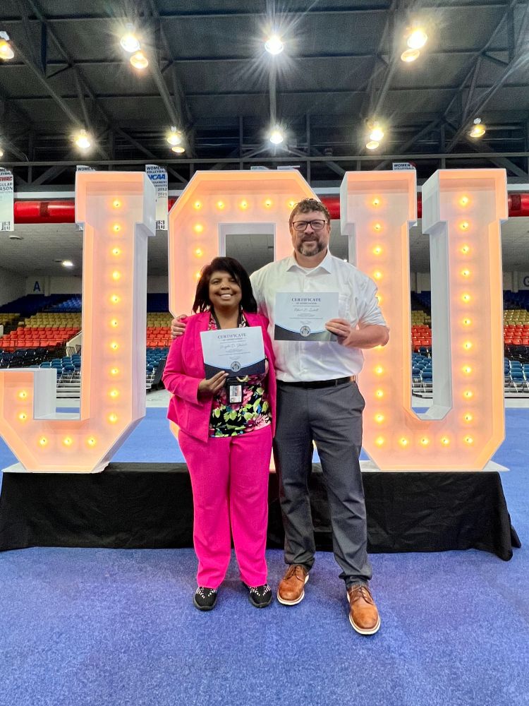 Two people in a large gymnasium with stands in the background pose in front of large white letters illuminated by light bulbs, spelling "JSU." They smile at the camera and hold up white and blue certificates.