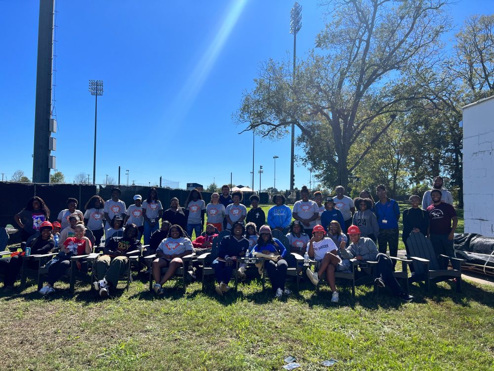 A large group of students, most of whom are wearing gray and orange Home Depot shirts, pose on lawn chairs outside on a green lawn. Trees and tall stadium lights are in the background.