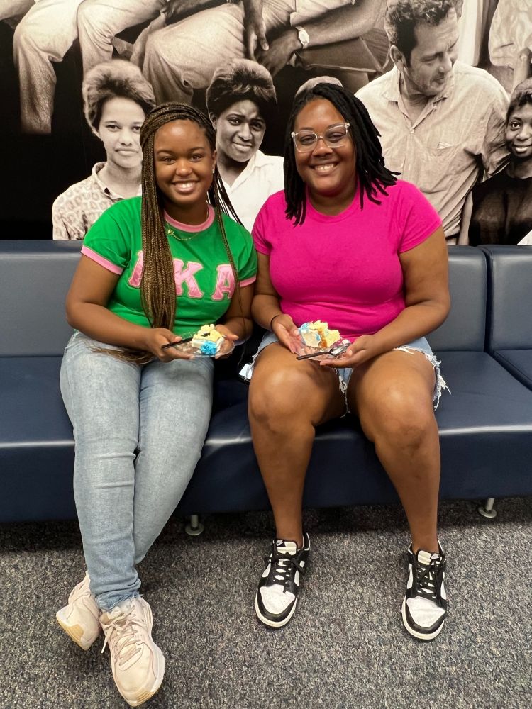 Two young women sit next to each other on a gray-blue couch in front of a photograph collage. They both hold slices of cake and smile at the camera.