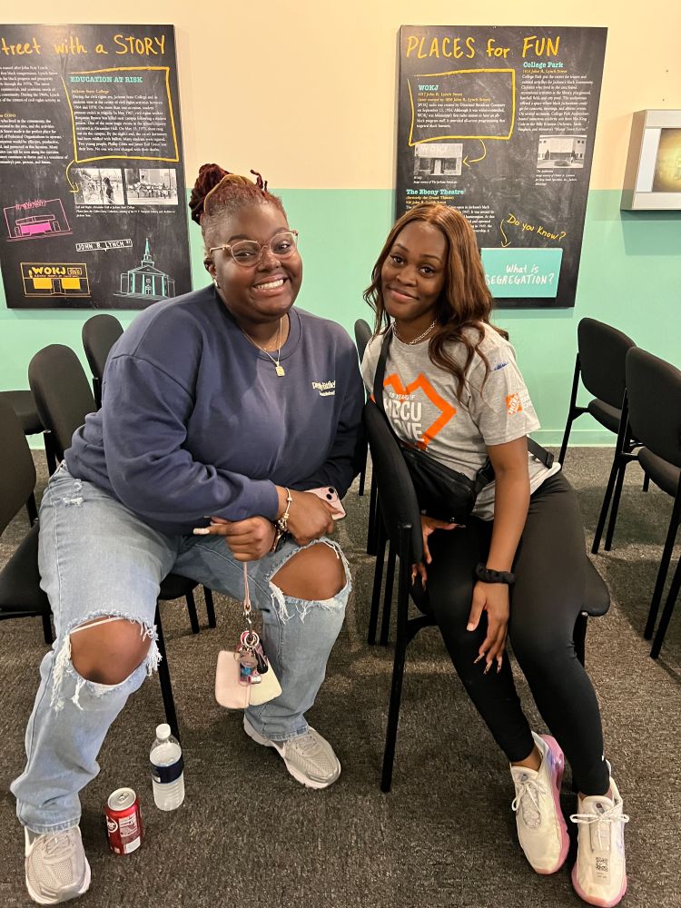 Two young women sit next to each other inside an exhibit room with informational wall posters with text and photos behind them.