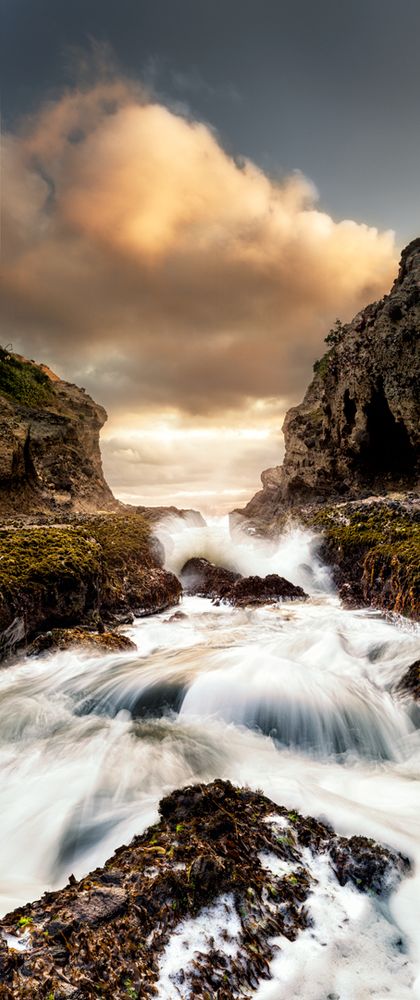 Water rushes through a channel in the rocks  at Kotau Point. Te Henga. In the foreground is a rock covered in barnacles. The sky is cloudy and the sun is low as it is just before sunset. 