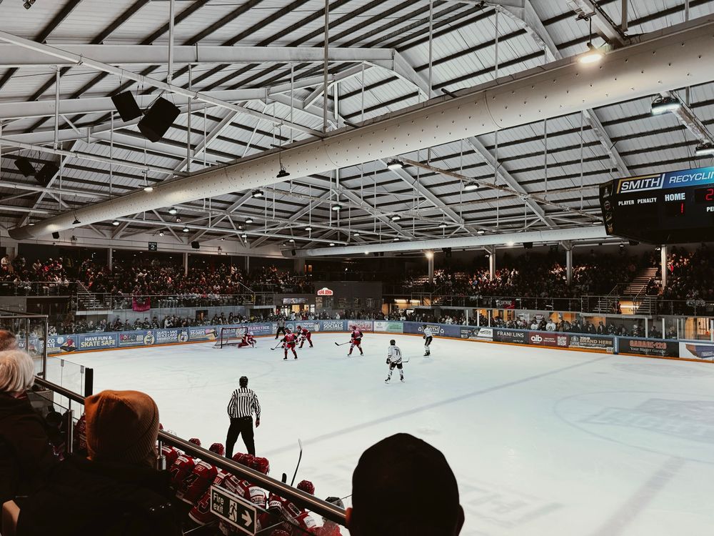 Live ice hockey game at a buzzing arena. Red and white teams face off as fans fill the stands. The scoreboard shows a 1–1 tie in the first period. Bright lights, a visible referee, and rink-side ads complete the energetic scene.