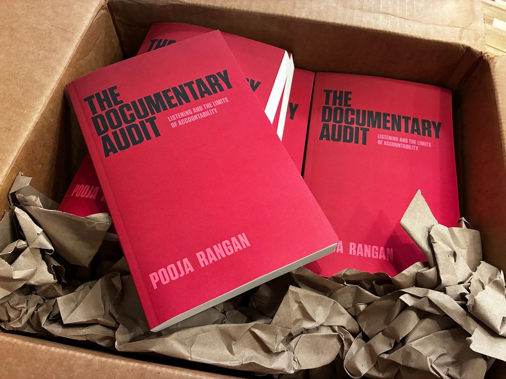 Copies of a book nestled in a cardboard box. Description of the book cover: The title of the book (The Documentary Audit) appears in bold black text against a deep crimson background that fades to pink toward the bottom right. The book's subtitle (Listening and the Limits of Accountability) and the author's name (Pooja Rangan) appear in smaller pink letters.