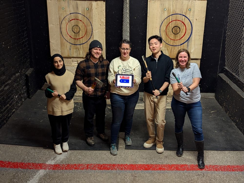 A group of 5 people holding axes posing in front of 2 axe throwing lanes