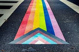 A progress pride flag painted onto a street crosswalk, with crosswalk intersection striping on the left hand side. 