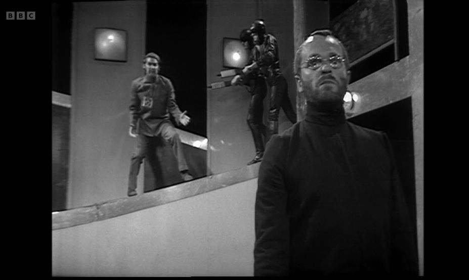 A screenshot from the eighth episode of the Doctor Who serial The War Games. On the right hand stand of the screen stands the War Lord (played by Philip Madoc) who appears to be looking either directly at, or just above, the camera. On the left side you can see a ramp behind him, rising from bottom left to top right. Halfway up the ramp stands the War Chief (Edward Brayshaw) who is shouting and gesturing towards the War Lord. At the top of the ramp stand two guards in entirely black outfits, pointing sci-fi guns at the War Chief.
