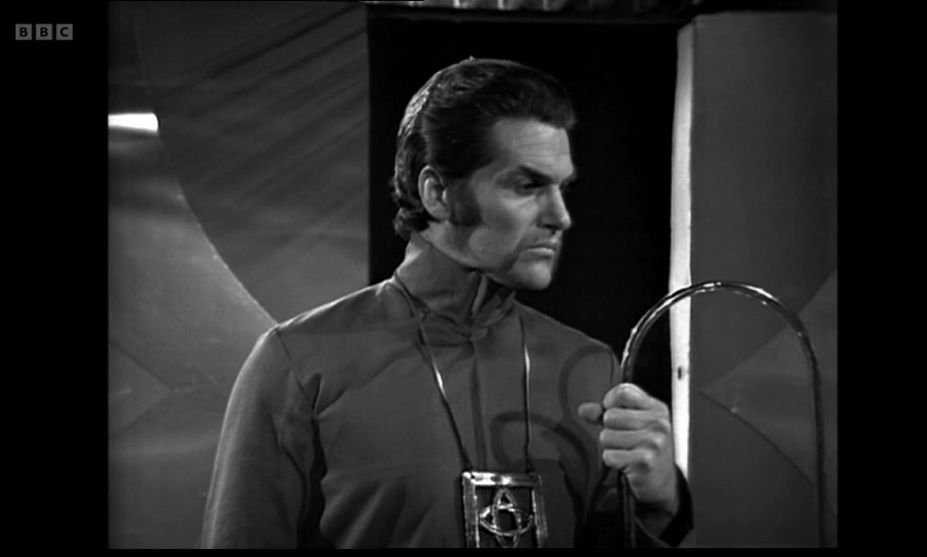 A still image from the third episode of the Doctor Who serial The War Games. Actor Edward Brayshaw is playing the War Chief. He is looking to the right while holding onto a metal loop with his left hand.