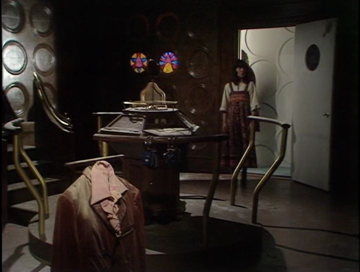 A screen shot from the first episode of the Doctor Who serial Masque of Mandragora. The image is inside the wood-panelled second control room. In the foreground a brown leather jacket and pink shirt are posed. Behind that is the new central console. Behind that, Sarah-Jane Smith stands in the doorway.