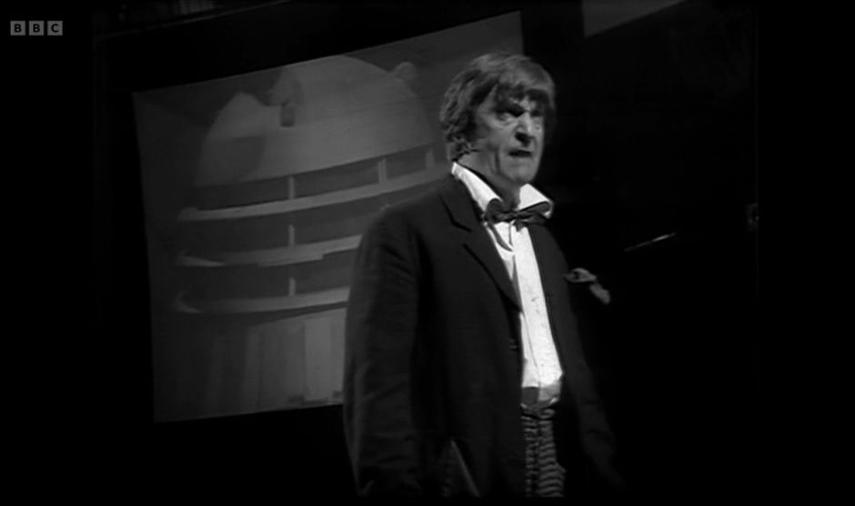 A screenshot from the Doctor Who serial The War Games. Just right of centre stands the Second Doctor (played by Patrick Troughton), addressing characters standing offscreen to the right. Behind him and slightly to the left is a square screen with a projected image of a dalek on it.