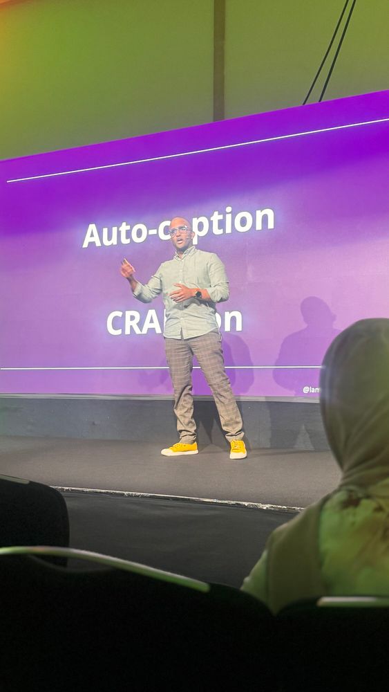 Ahmed on stage speaking to an audience, wearing green shirt, patterned brown trousers and yellow shoes. He is standing in front of the screen.