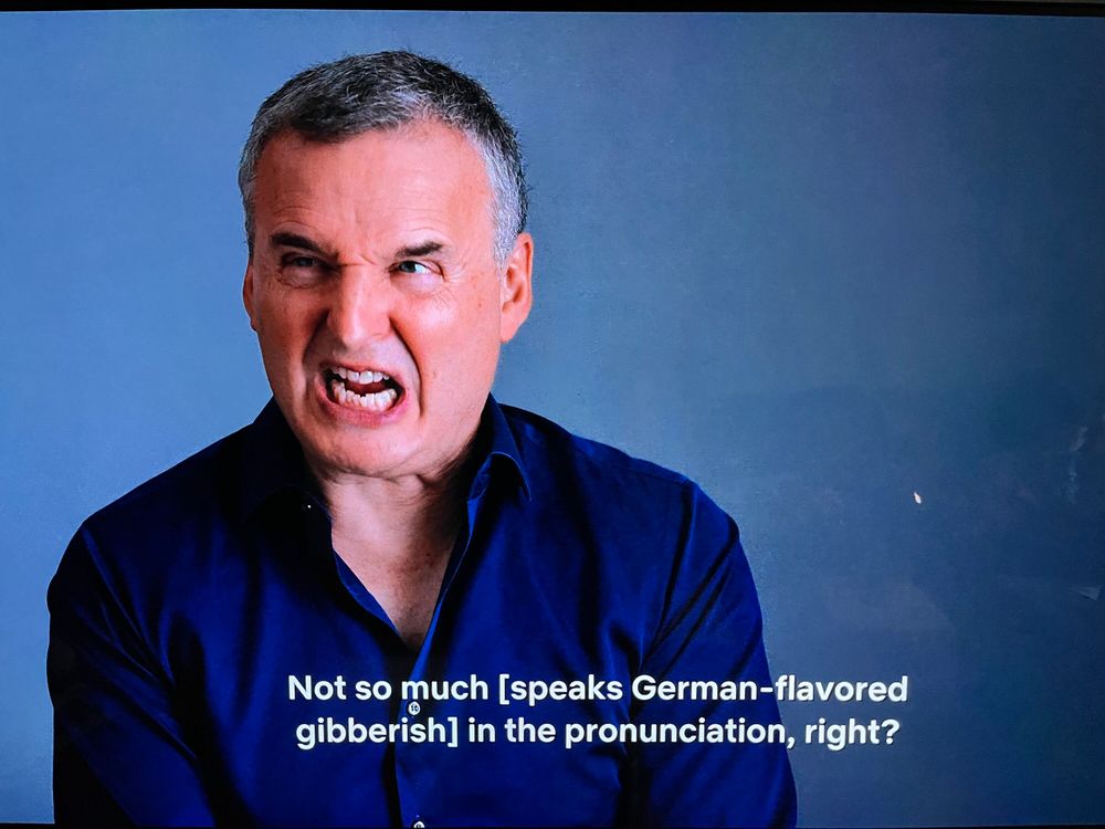 A still in an episode of 'Somebody Feed Phil' where Phil Rosenthal is making a facial expression that implies he is trying. The caption reads "Not so much [speaks German-flavored gibberish] in the pronounciation, right?