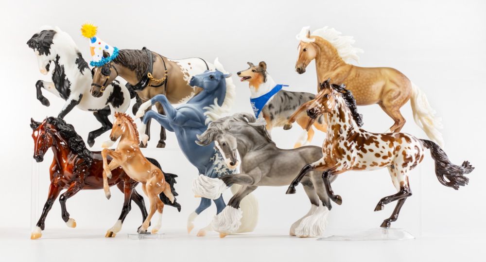 Photo of all 8 revealed BreyerFest 2025 Special Run models, lined up in 2 rows against a white background. The secret "Surprise" as always is not included. The photo is taken by Breyer at their HQ. Top L-R: Get This Party Started (Black Pinto Croi), Hats Off To You (Dun Appaloosa Old Timer), Life's a Journey (Blue Merle Collie), Future is Bright (Palomino Bandalero). Bottom L-R: Good Times & Great Friends (Bay Mustang Mare & Chestnut Foal), To The Ties That Bind Us (Blue Deco w/Horse Silhouette Appy Pattern Silver), YOLO (Dapple Gray Brishen), and Let The Good Times Roll (Buckskin Appaloosa Show Jumper). The models (except the dog) are 1:9 scale (the dog is physically smaller than the horses but is larger than them in terms of scale). 