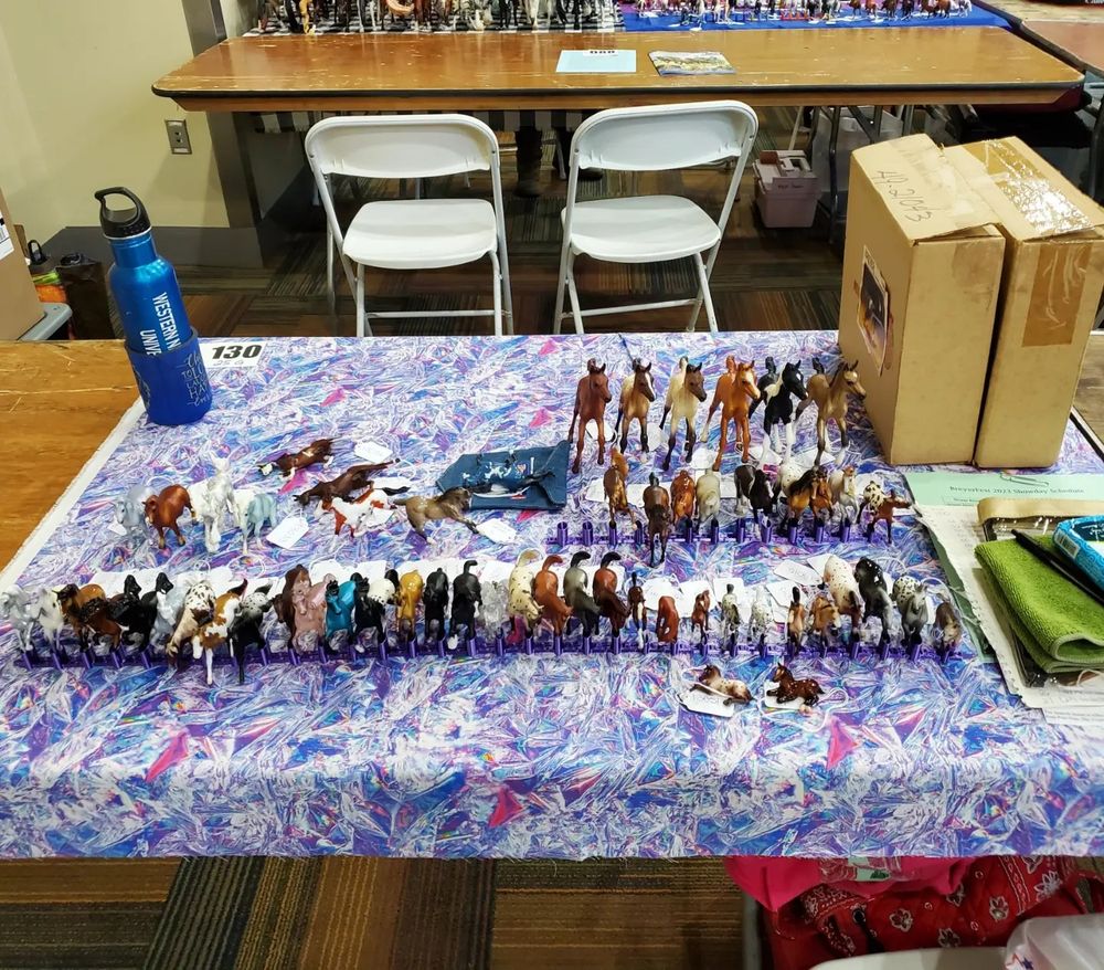 A photo of Breyer Model Horses on a wooden banquet table covered by a colorful cloth. There are mostly Stablemate scale models (1:32) held up from falling by metallic purple Stable Snaps (a 3-D printed product designed by my boyfriend). There are a few larger models towards the back - Classic scale (1:12) foals. I also have a blue water bottle, a couple of cardboard boxes containing more Breyers, and miscellaneous items on the table. The tables of other show entrants are visible in the background. Photo taken during BreyerFest Live July 2023. 