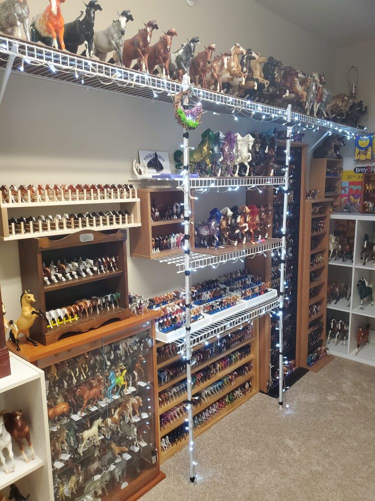 A collection of neatly arranged model horses of various sizes, shapes, and colors on a few different shelves. There is a string of LED lighting woven through the rack-style shelving unit which is illuminated for the picture.