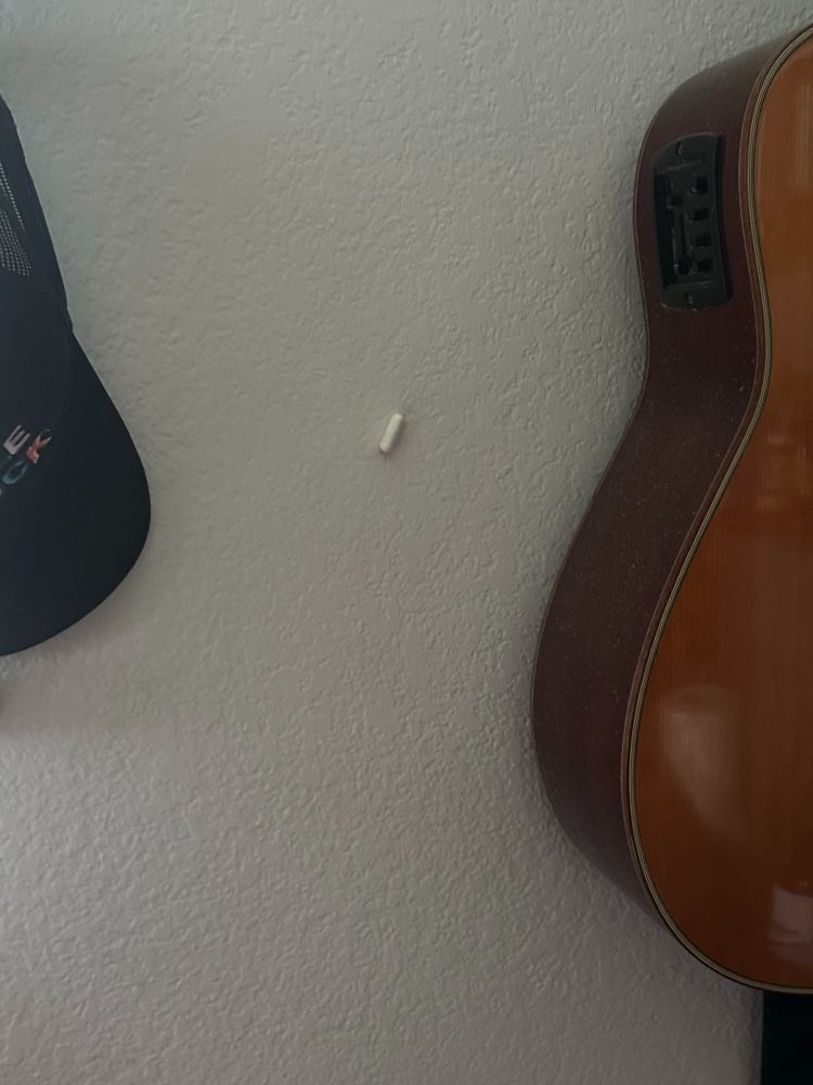 an oblong white gel-pill supplement, approximately .75 inches in size, adhered to a textured wall, in between an acoustic guitar and a hanging black hat after a laughter-induced spit take fired it across a room. The poster brands this act a Spit Pill Missile™️